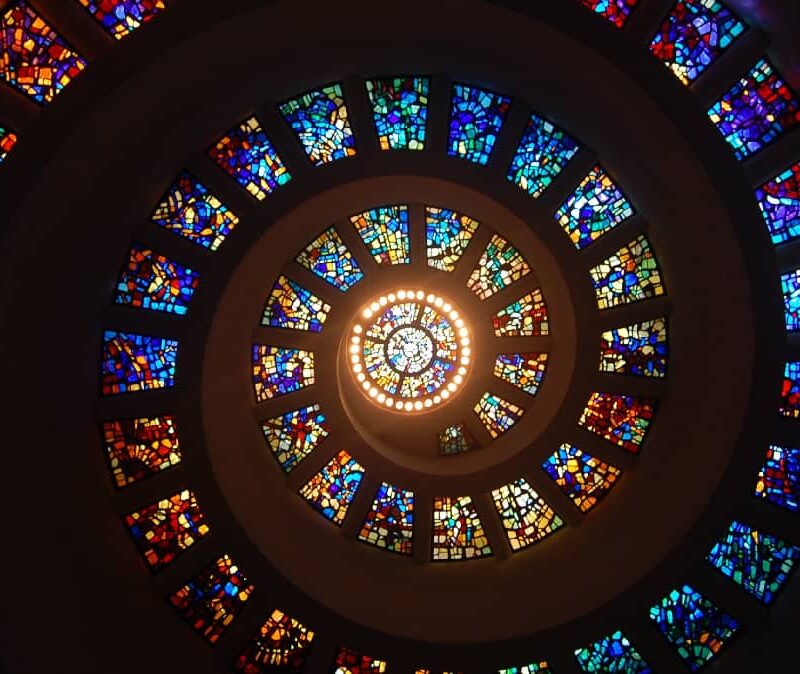 A spiral staircase with vibrant stained glass windows, symbolizing growth and embodying the theme 'progress, not perfection'.
